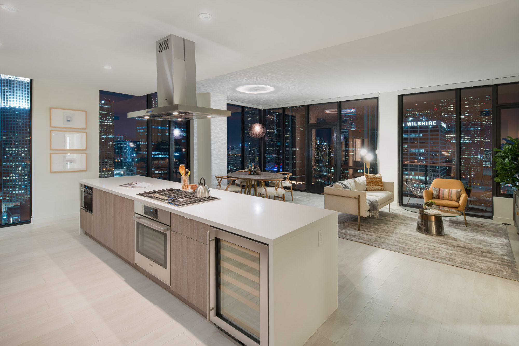 A luxurious open floor plan kitchen island facing a living room, surrounded by floor to ceiling glass walls with evening city views of downtown LA