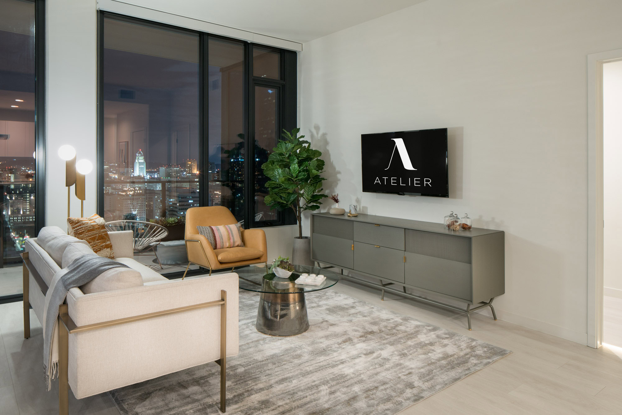 A living room in Ateliers luxury apartment in downtown Los Angeles CA with floor to ceiling windows showing evening city views