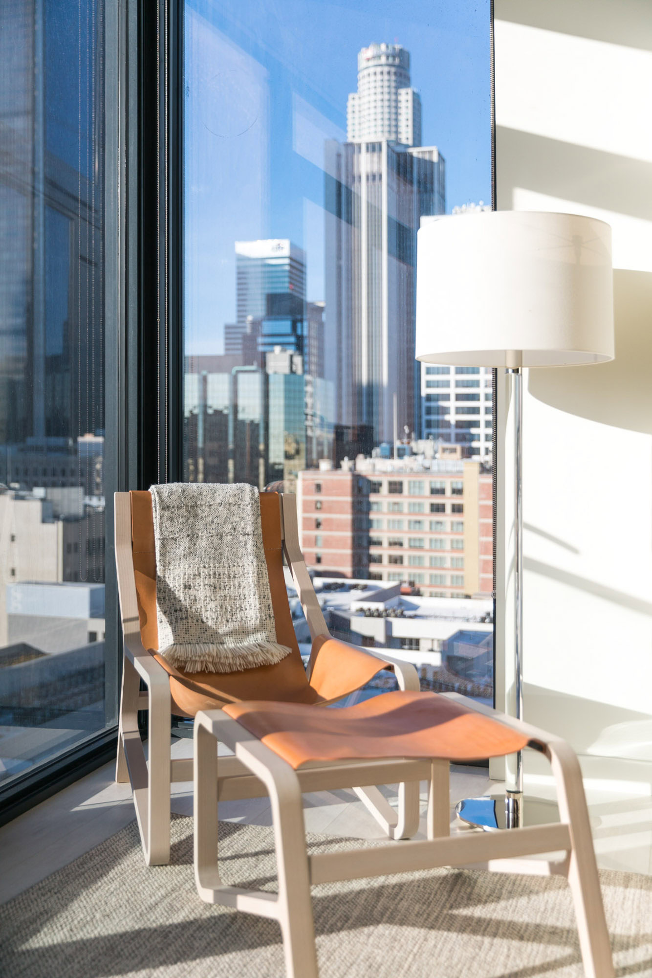 A living room reading corner with glass floor to ceiling windows in the background showing city views at Ateliers DTLA Apartments