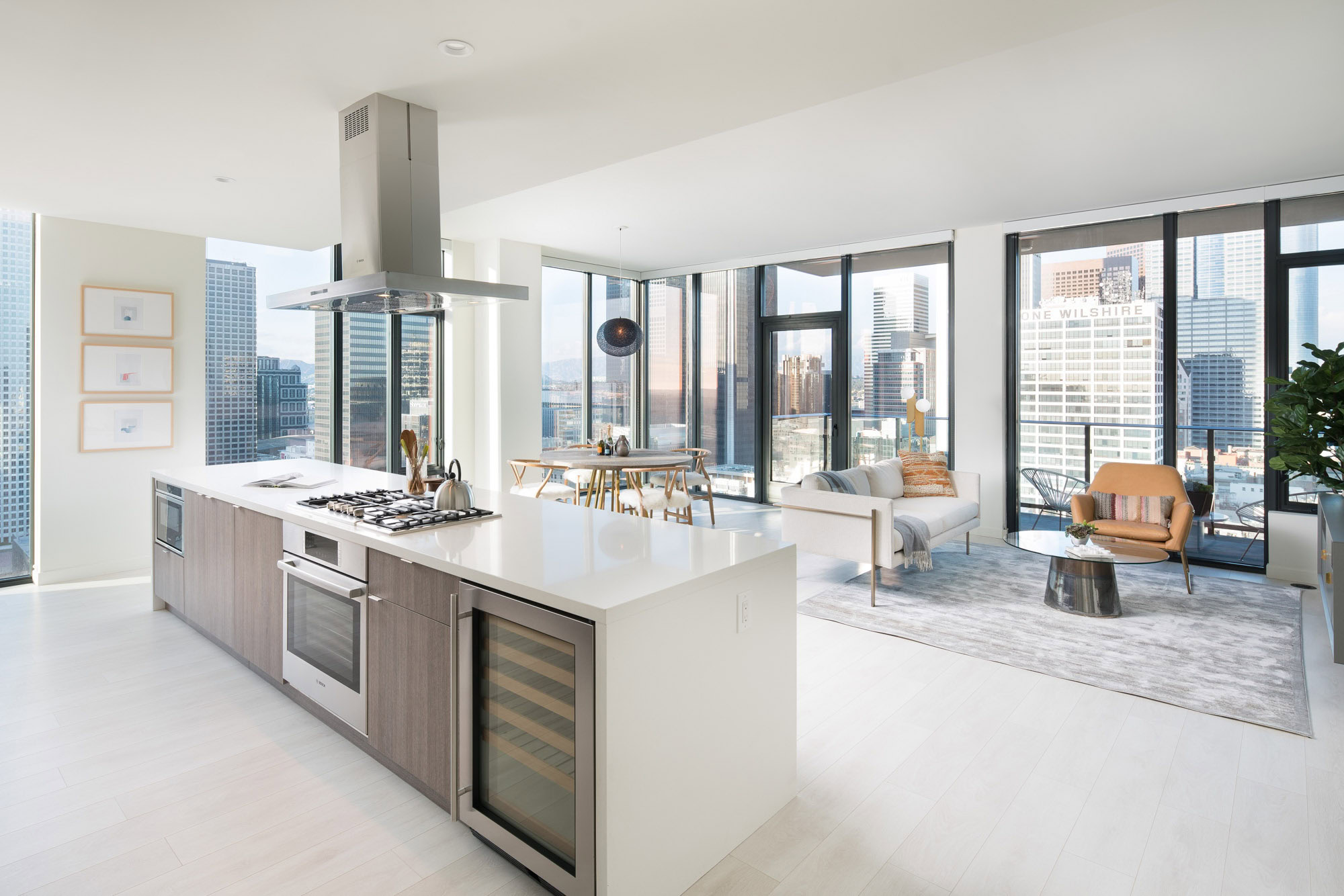 A luxurious open floor plan kitchen island facing a living room, surrounded by floor to ceiling glass walls with daytime city views of downtown LA