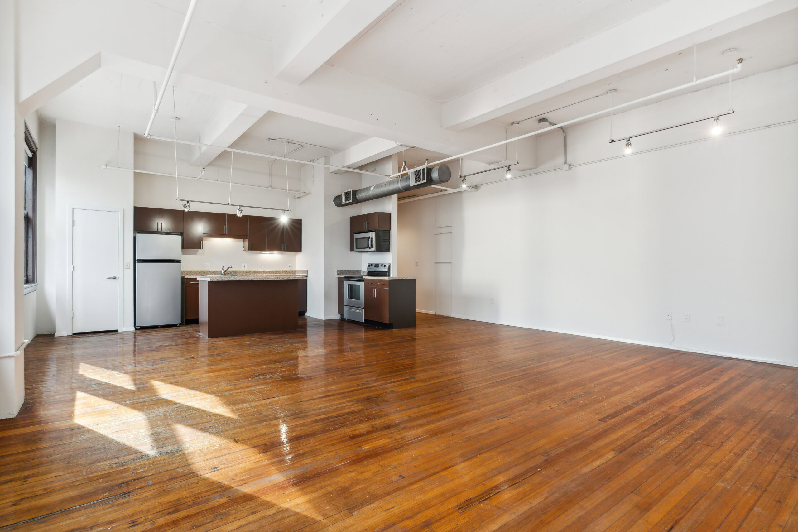 Open plan living and dining room area with natural lighting, polished wood flooring and exposed piping in a vacant boutique apartment at The Wilson in Dallas Texas.