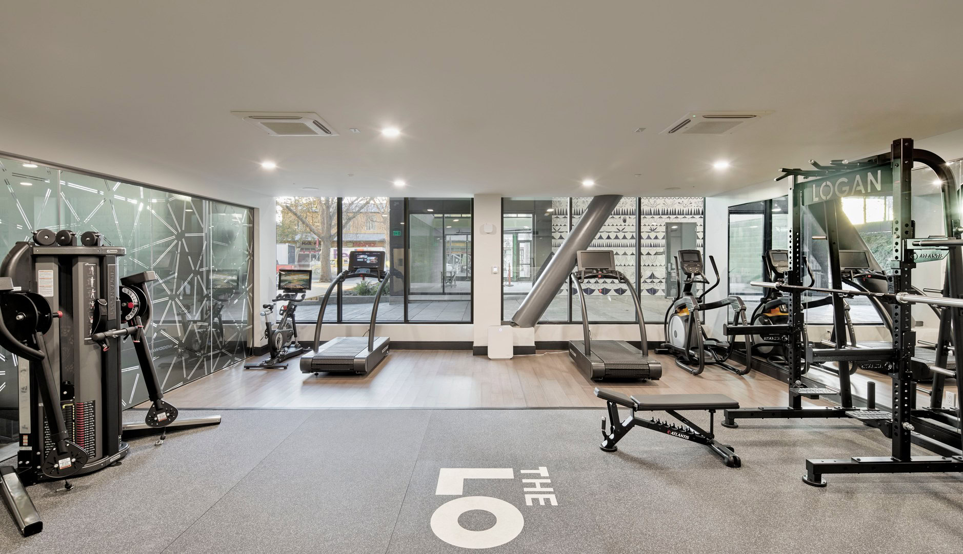 A modern fitness center with cable machines, treadmills, an elliptical and stationary bike machines facing a window with street views at The Logan in Temescal, Oakland CA