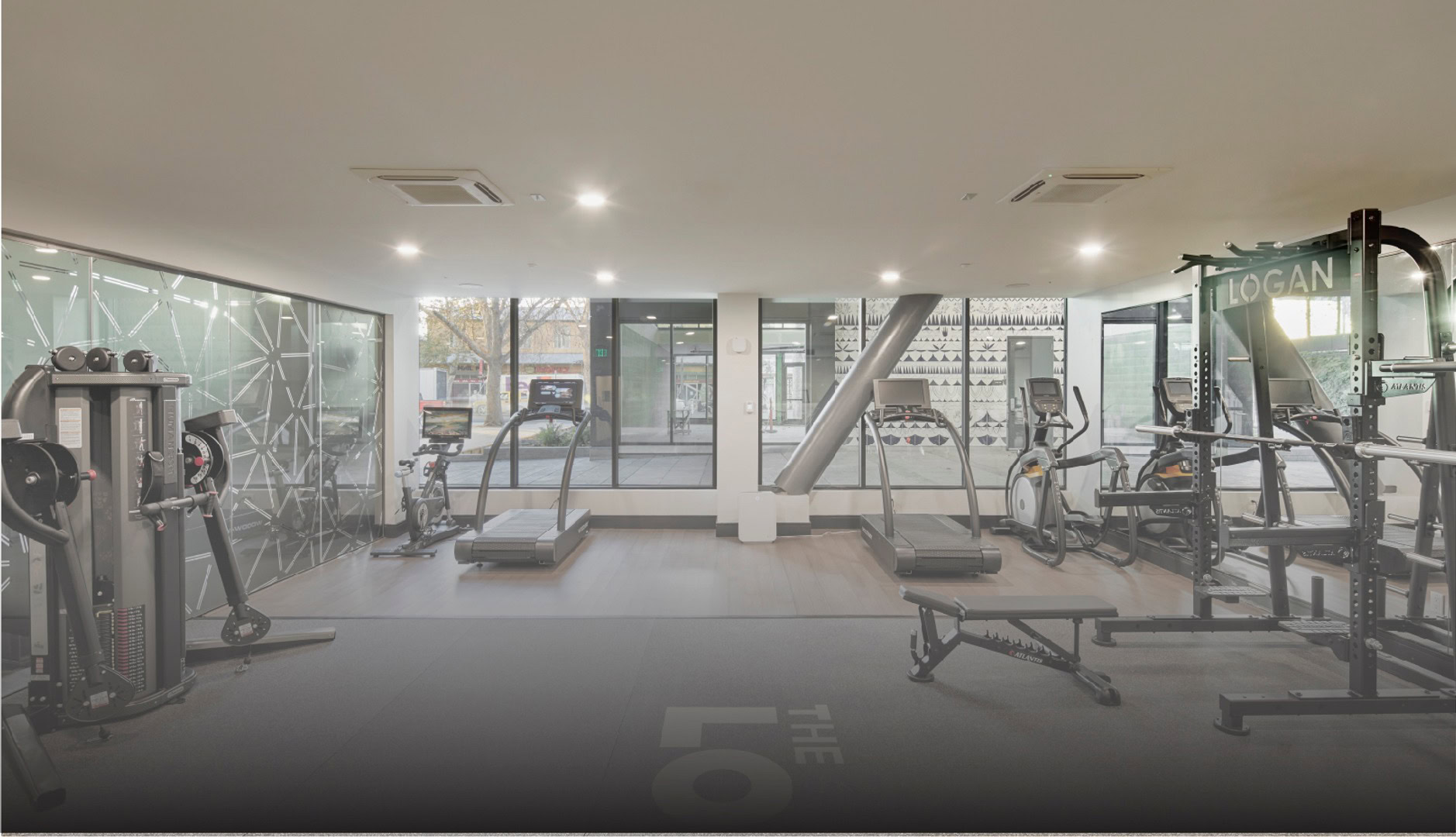 A modern fitness center with cable machines, treadmills, an elliptical and stationary bike machines facing a window with street views at The Logan in Temescal, Oakland CA