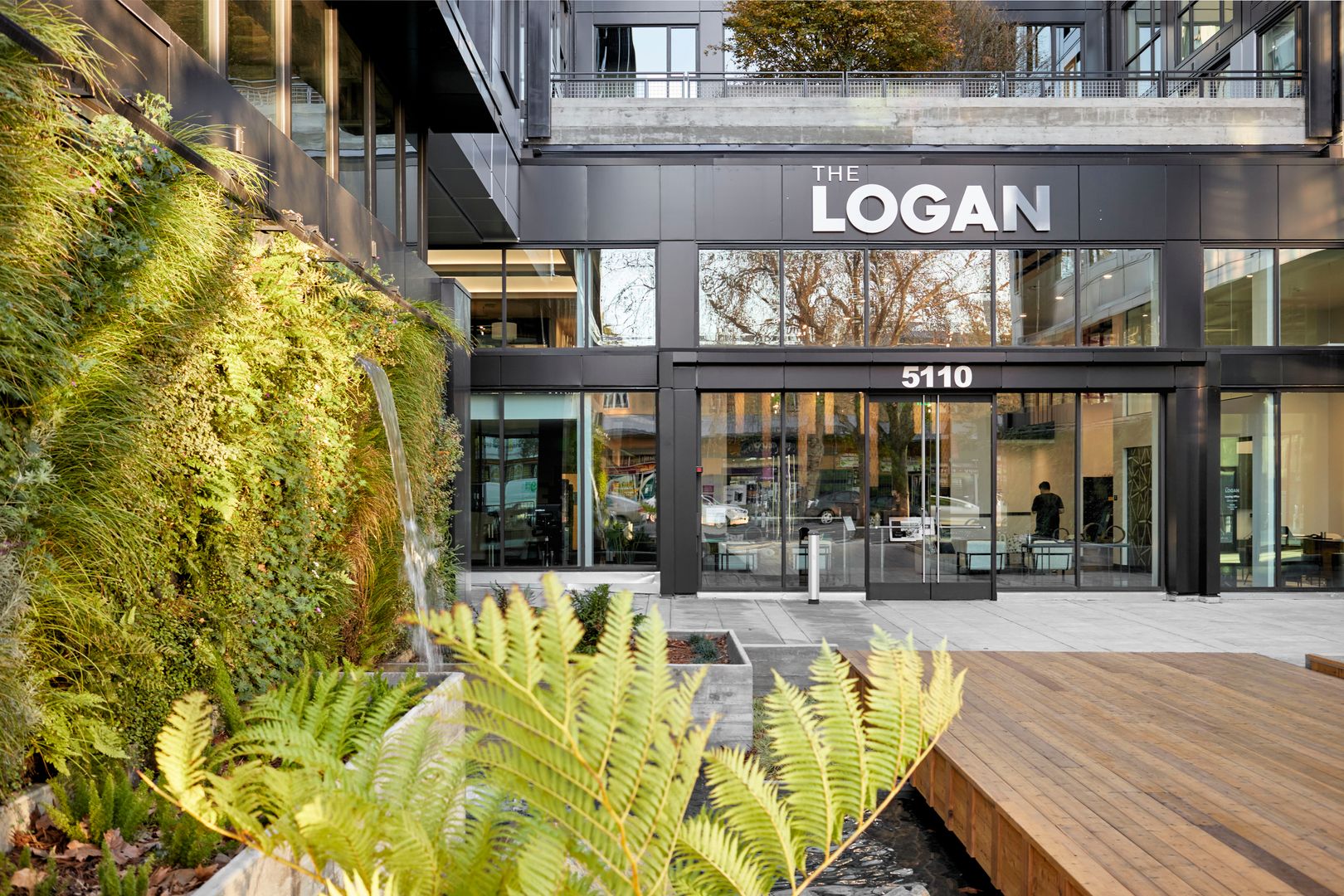 An entrance with biophilic elements including a wooden bridge surrounded by greenery and water feature in Temescal apartments, The Logan