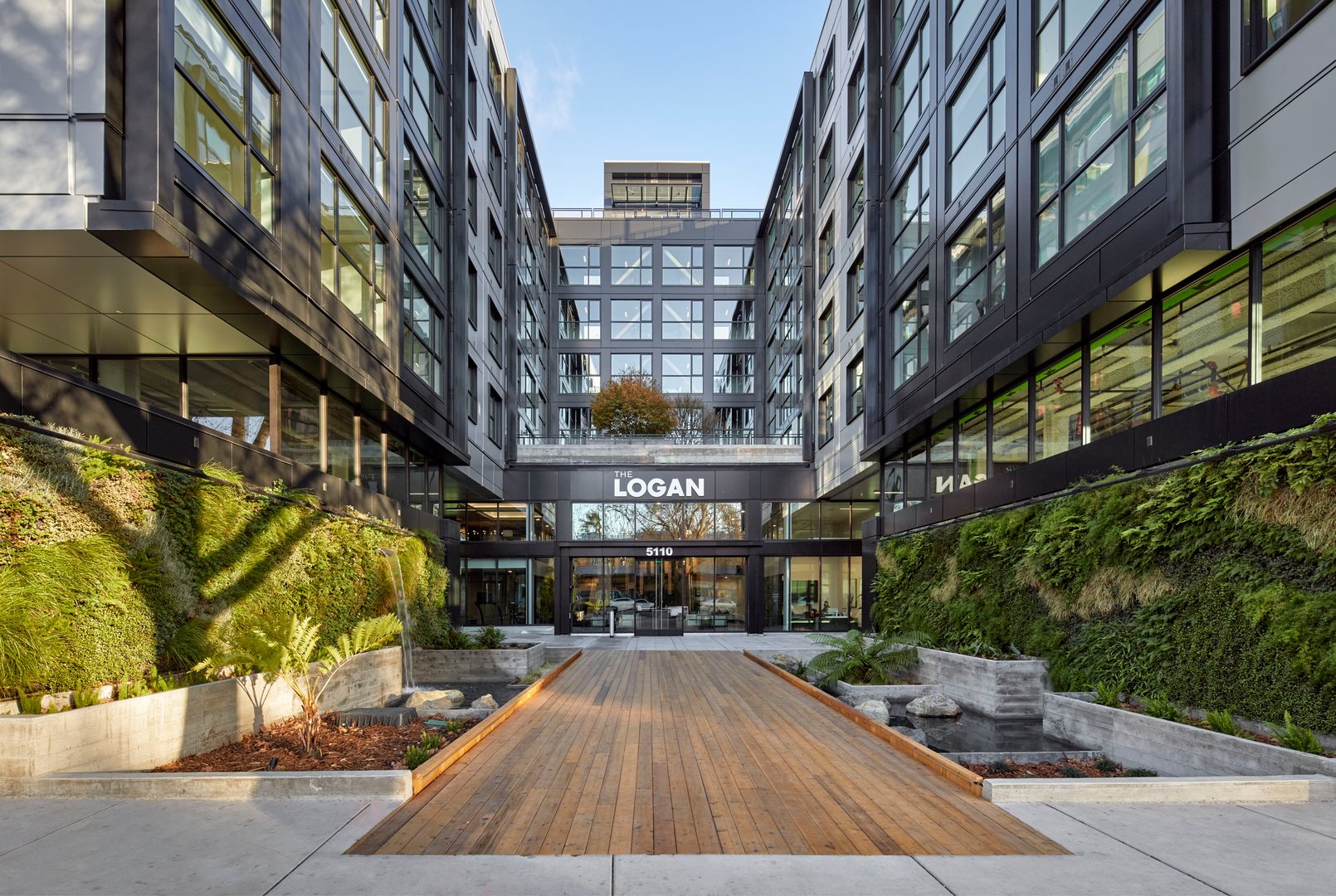 An entrance with biophilic elements including a wooden bridge surrounded by greenery and water feature in Oakland apartments, The Logan