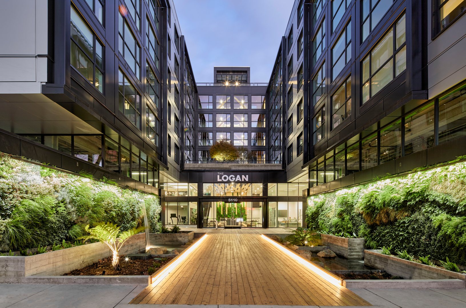 An entrance with biophilic elements including a lit up wooden bridge, greenery and a water feature in Oakland apartments, The Logan