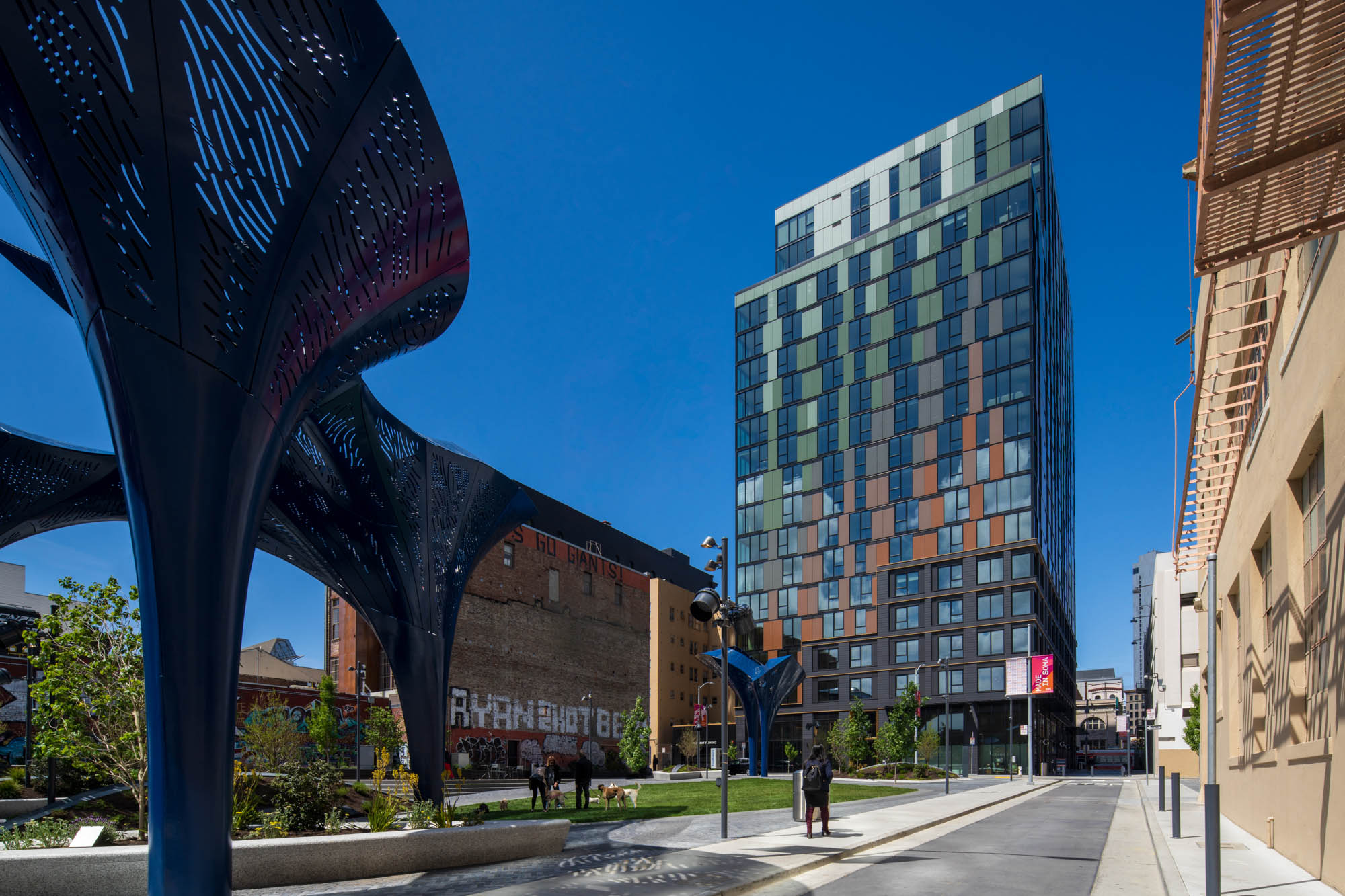 A modern urban scene with a tall, colorful glass building on the right under a clear blue sky. On the left, abstract dark sculptures adorn a green park area. The street has a calm, orderly vibe.
