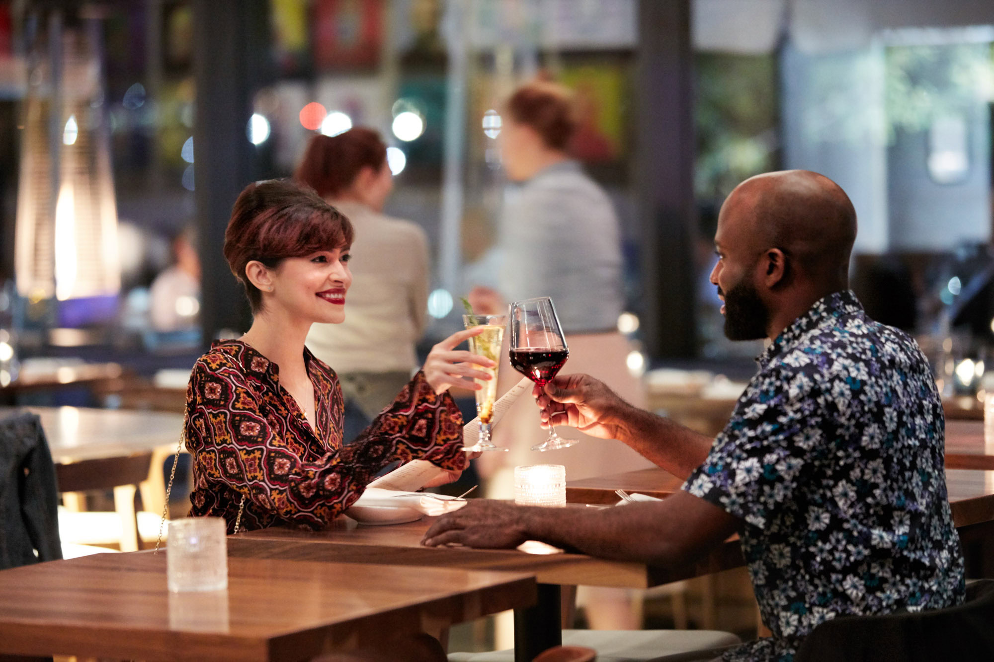 A couple sits in a cozy, dimly-lit restaurant clinking wine glasses and smiling warmly at each other. The atmosphere is intimate and joyful.
