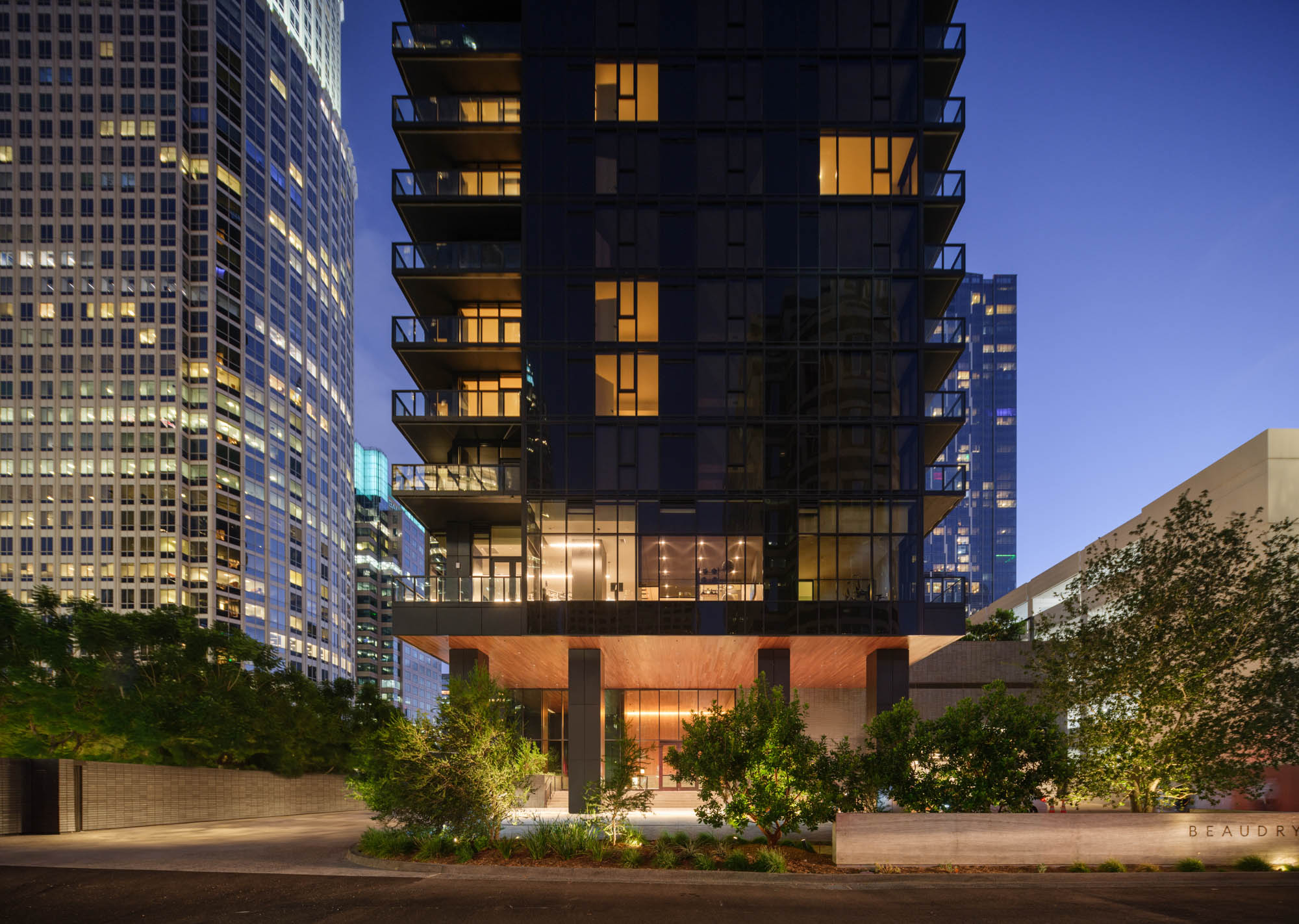 A modern high-rise building at dusk with glowing warm lights in windows, surrounded by greenery. Nearby, other illuminated skyscrapers rise into a clear evening sky.