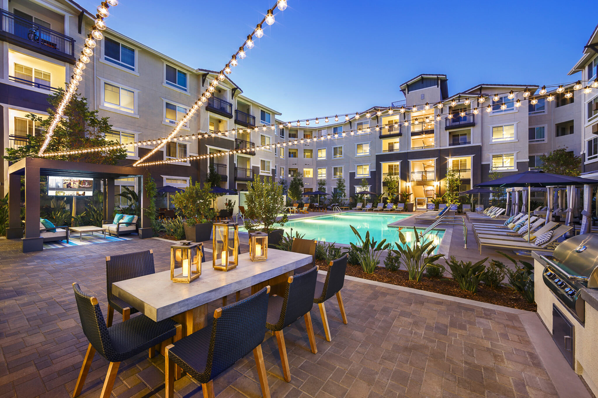 Luxury apartment courtyard at dusk with string lights, a modern outdoor dining table, lounge chairs, pool, barbecue area, and lush landscaping surrounded by multi-story buildings.