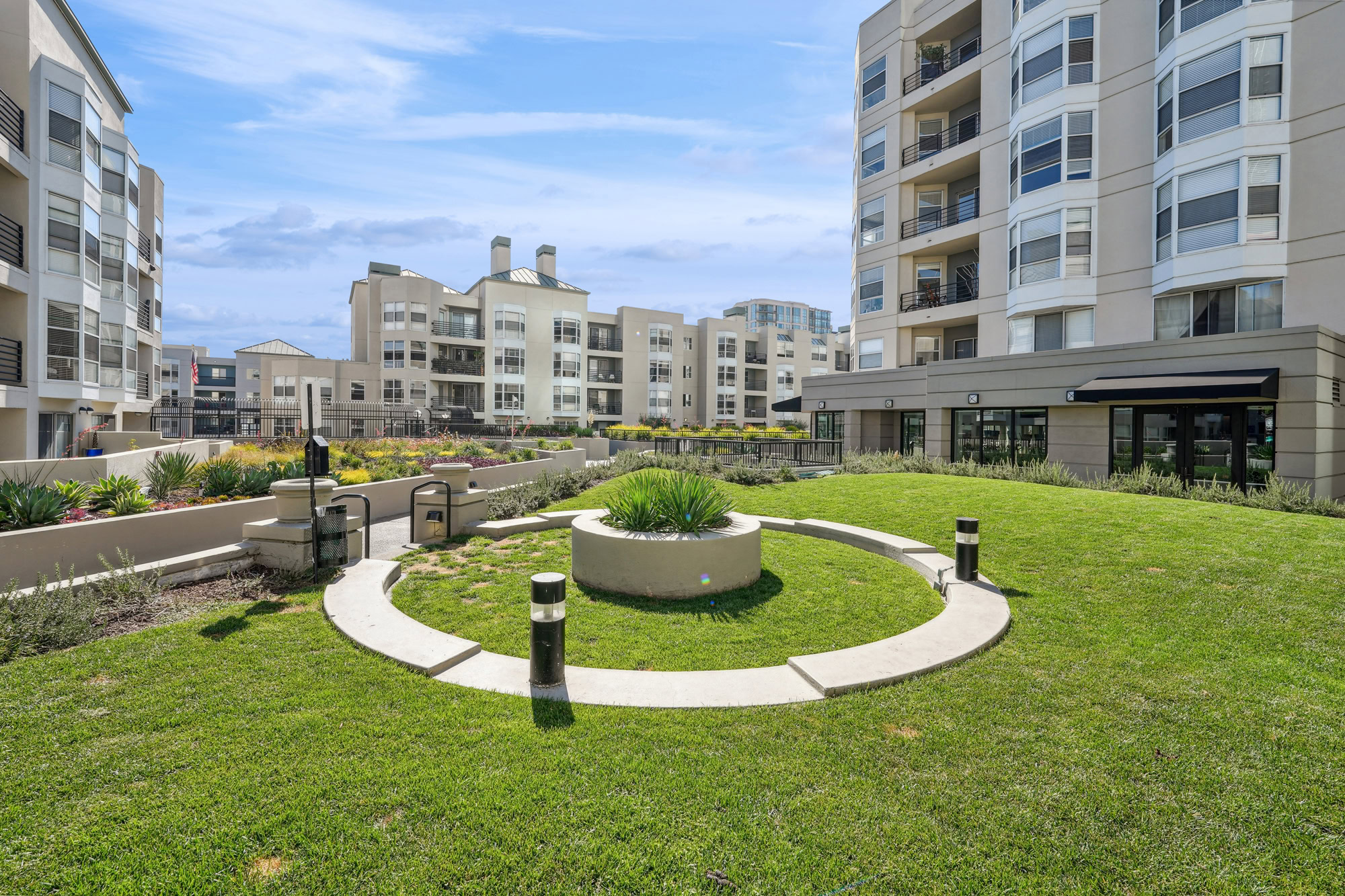 Modern apartment complex courtyard with manicured lawn, circular pathway, central planter, and surrounding multi-story buildings under a clear blue sky.