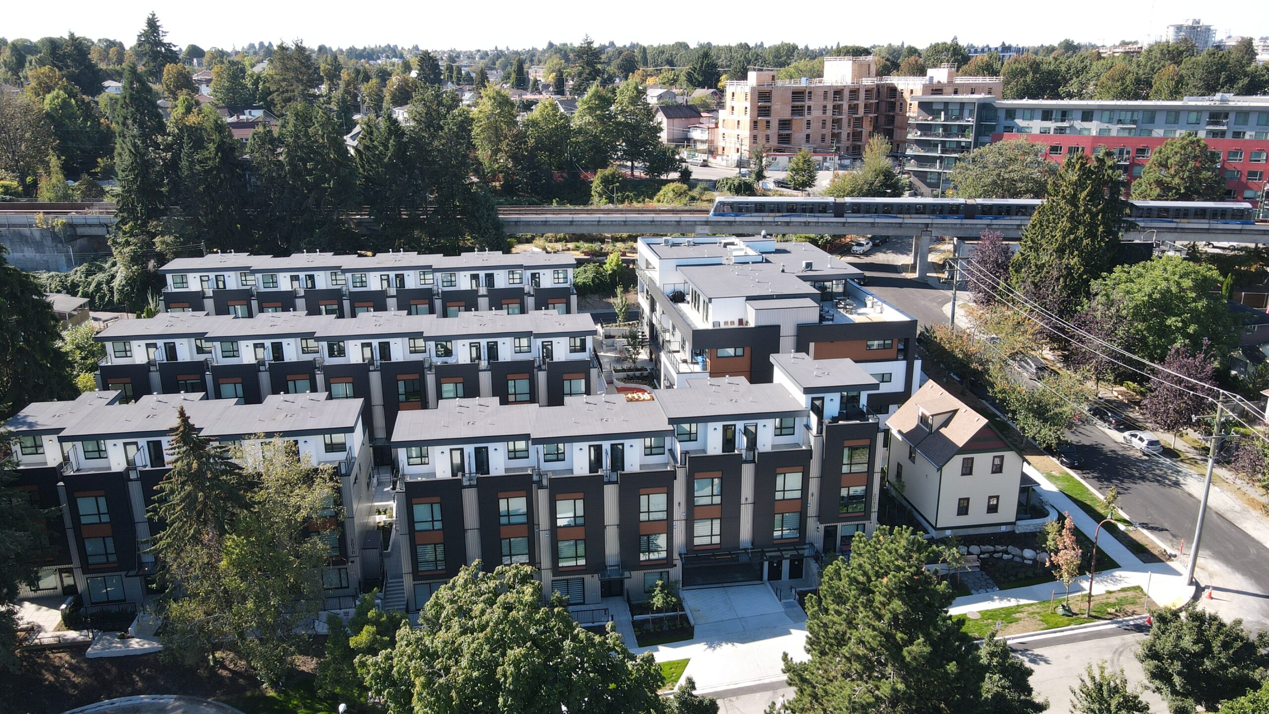 Aerial view of a modern townhouse complex surrounded by trees, with a train passing by on elevated tracks and mid-rise buildings in the background under a clear sky.