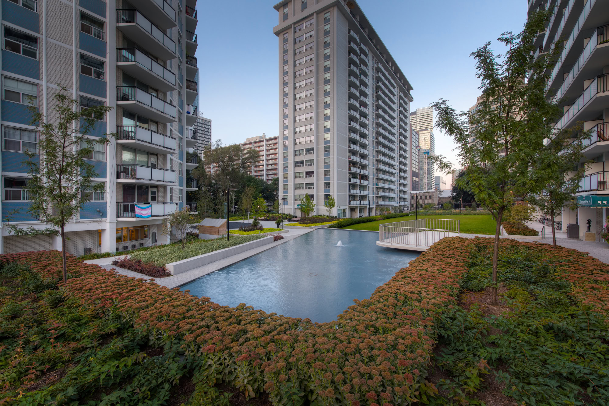 Urban scene with modern high-rise buildings surrounding a serene courtyard featuring a reflective pond, lush greenery, and peaceful atmosphere.