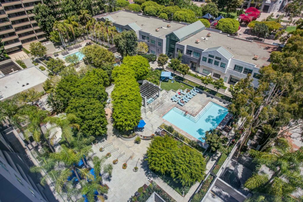 Aerial view of a lush courtyard with two swimming pools surrounded by trees, lounge chairs, and umbrellas between modern buildings on a sunny day.
