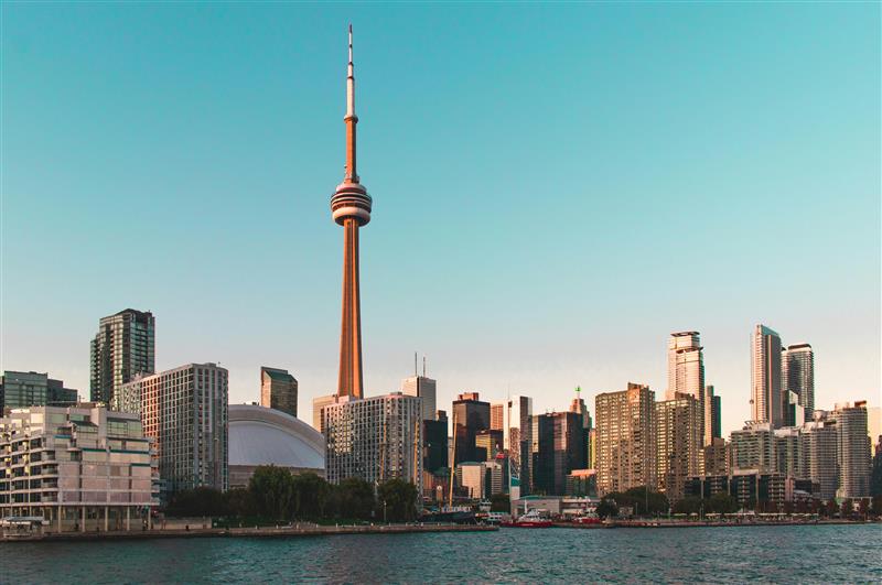 Toronto skyline at dusk with the CN Tower prominently visible among modern skyscrapers, set against a clear sky and reflected in calm water.