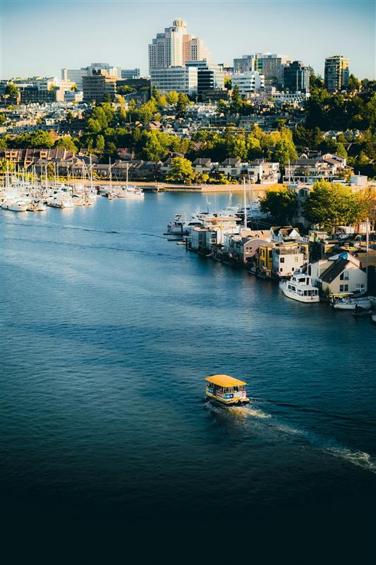 A small yellow boat travels on a river lined with houseboats and yachts, with a cityscape of tall buildings and green trees in the background under clear blue skies.