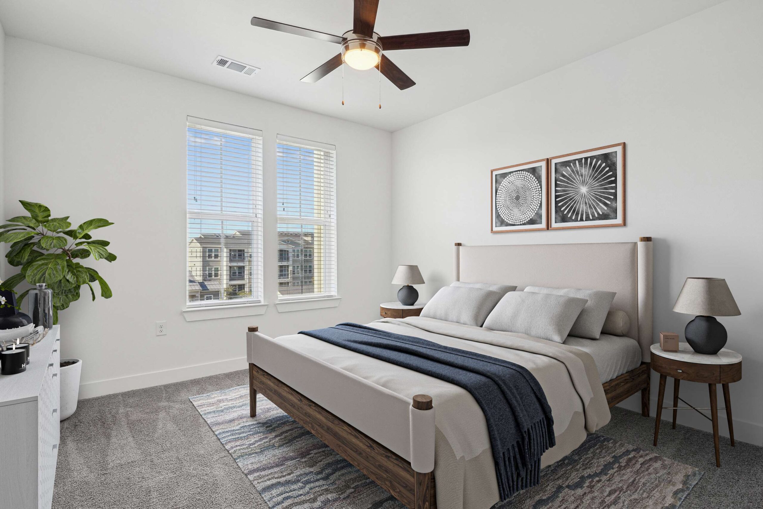 A serene bedroom with a modern style. Features a large bed with a beige headboard, blue throw, two side tables, lamps, plant, and geometric artwork on a white wall.