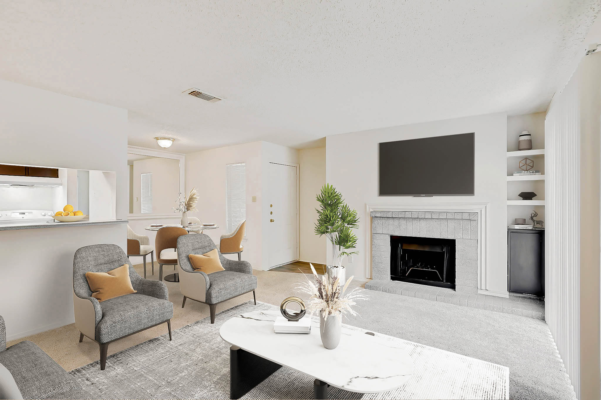 Bright living room with gray chairs, a white coffee table, a fireplace with a TV above it, and built-in shelves. A dining area with a table and chairs is visible near an open kitchen and front door.